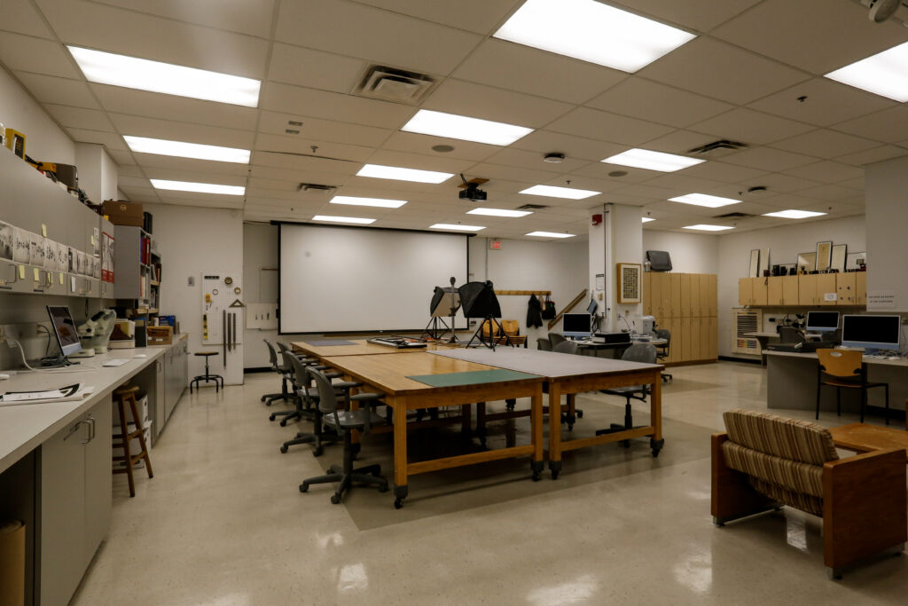 An interior view of a contemporary design or art studio classroom, featuring multiple large wooden work tables, computer workstations along the perimeter, a projection screen, and photography equipment like softboxes.
