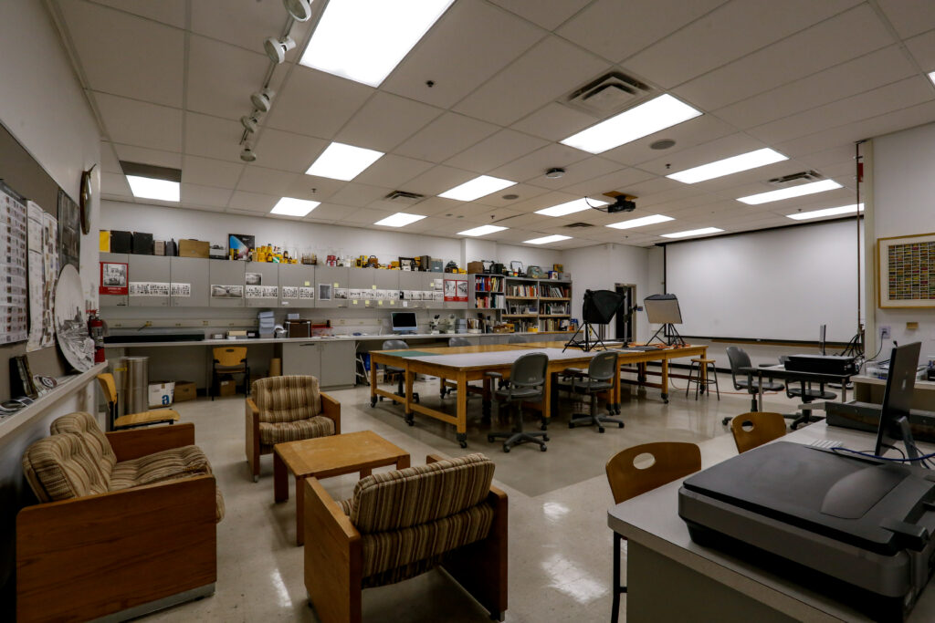 A spacious creative studio, possibly for photography or design, featuring workbenches, large communal tables, a small lounge area, photography lighting equipment, and a projection screen.