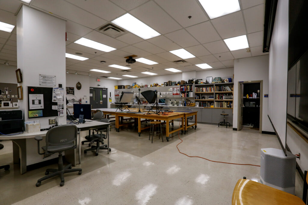 An empty college art and photography studio, featuring long wooden worktables with studio lighting setups, multiple computer workstations, and shelves stocked with art supplies and books under bright overhead lights.