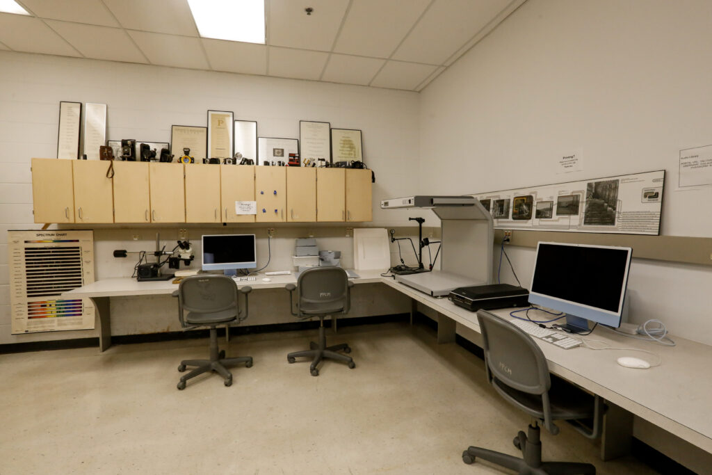 An academic or professional photo and digitization lab featuring two computer workstations, a microscope station, a large overhead book scanner, and a collection of antique cameras displayed on overhead cabinets, suggesting a space for visual arts or archival work.