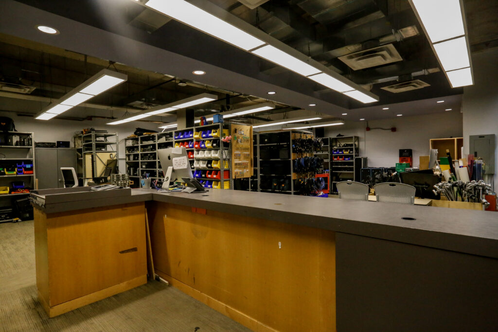 A media equipment rental or checkout desk with an iMac, phone, and shelves overflowing with cameras, cables, and production gear in a large, brightly lit room.