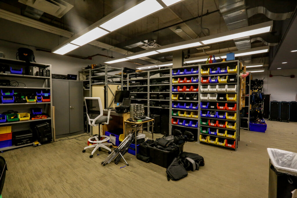 A well-lit, industrial-style equipment room featuring numerous metal shelving units filled with electronic devices, tools in colorful bins, a central workstation with a computer, and large rolling cases, suggesting a technical inventory or workshop.