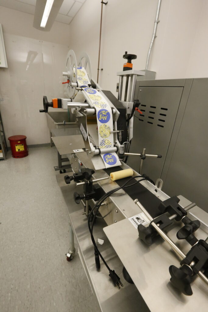 An industrial labeling machine in a manufacturing setting, feeding a roll of circular 'Get Inked' stickers onto a conveyor belt for application.
