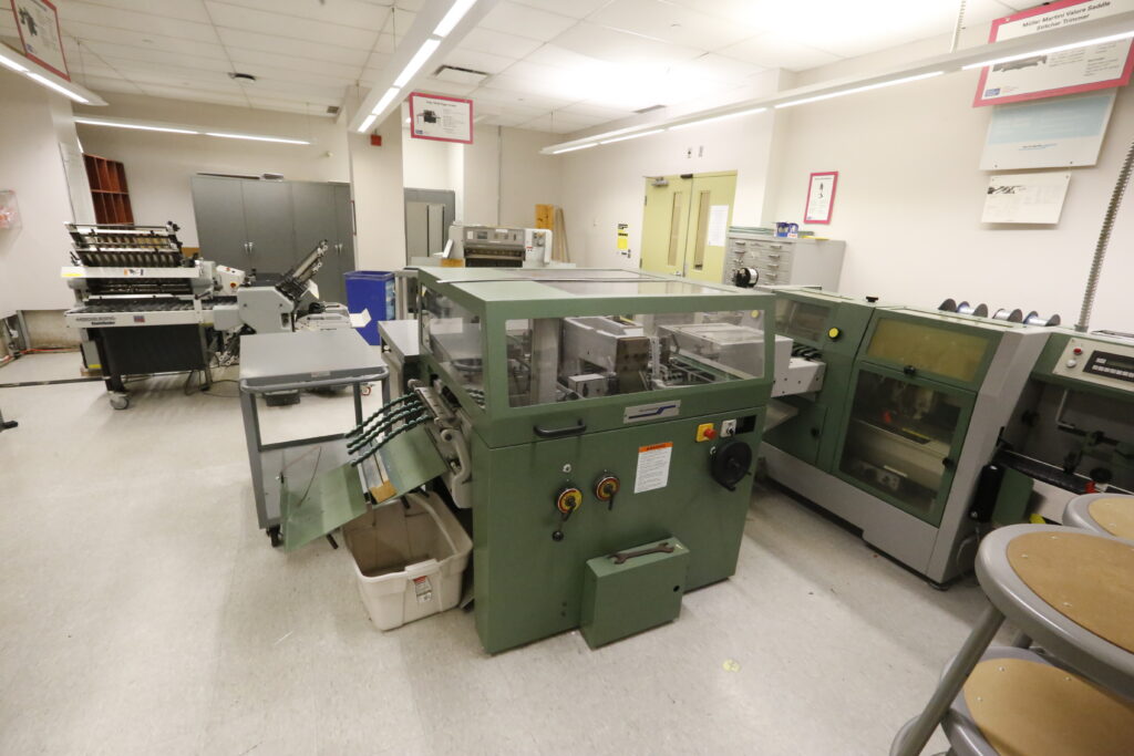 An industrial bindery equipped with a dark green Müller Martini saddle stitcher, a white Heidelberg Stahlfolder, and a grey paper cutter, set in a brightly lit room with informational signs and storage cabinets.