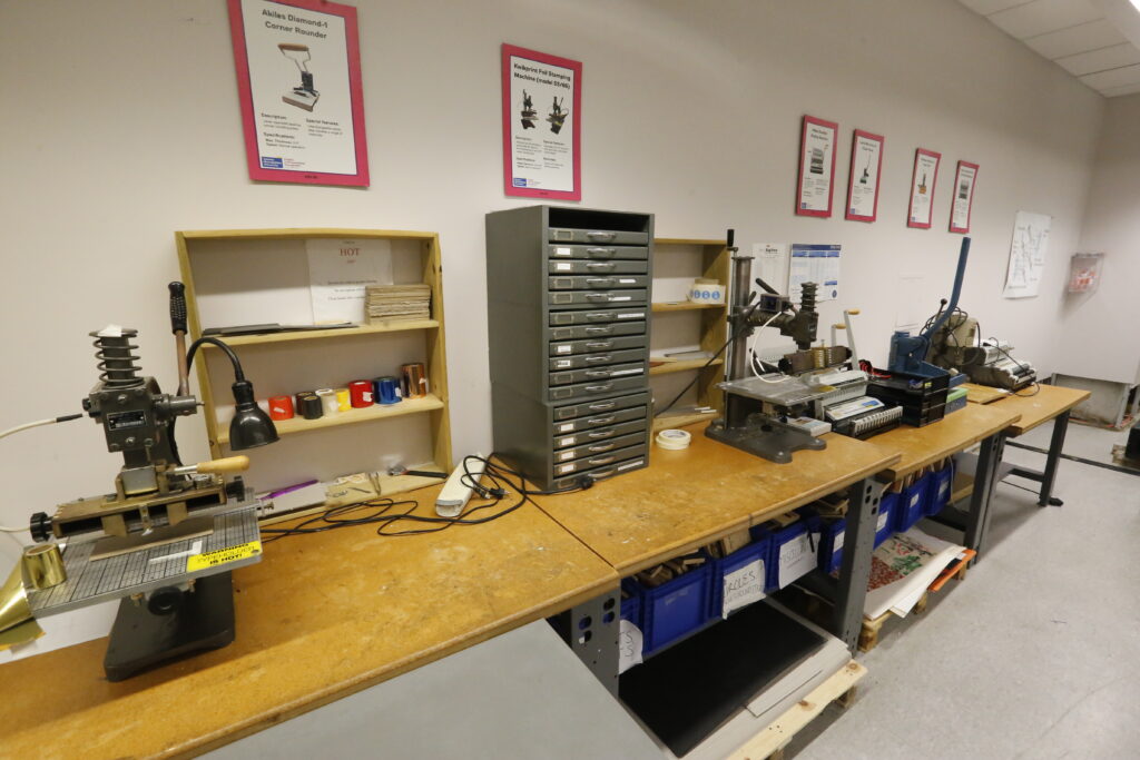 An organized workshop with several specialized machines, including a hot stamping press and binding equipment, on wooden workbenches, with informational posters on the wall.