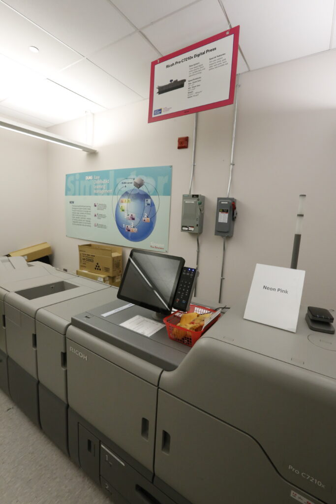 A Ricoh Pro C7210x digital press with a touchscreen interface, a 'Neon Pink' sample sign, and an overhead poster detailing the press's advanced color capabilities, including its 5th color station.