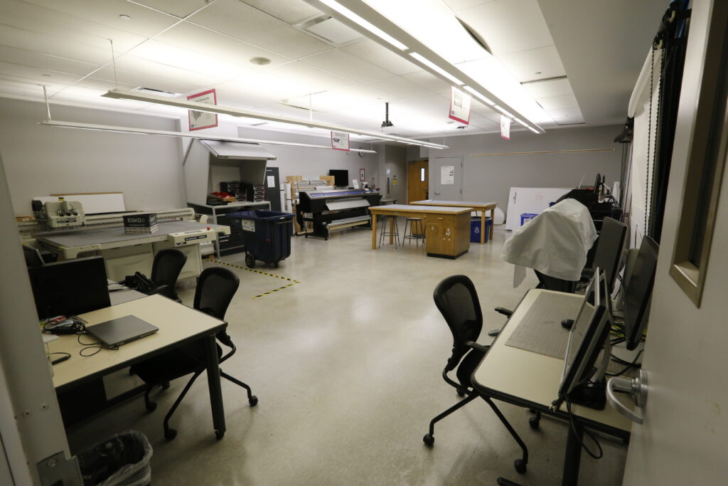 An organized fabrication lab or print shop, featuring a large Esko Kongsberg cutting table, a wide format printer, and multiple computer workstations with office chairs on a light gray floor.