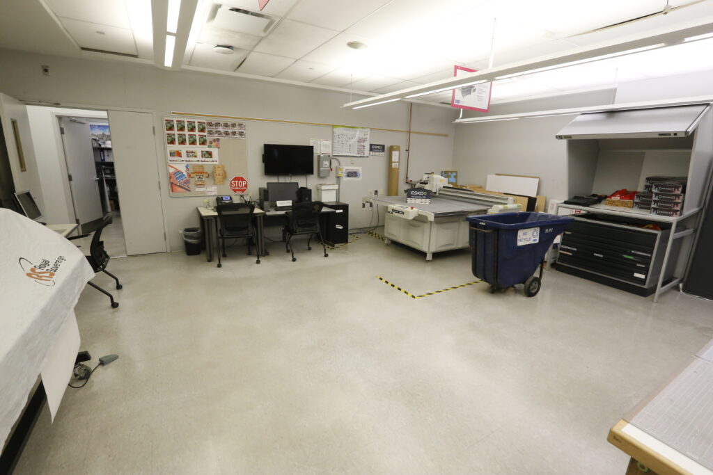 An organized design and fabrication workshop featuring an Esko Kongsberg V20 flatbed cutter, multiple computer workstations, and shelving for supplies, suggesting a creative production environment.