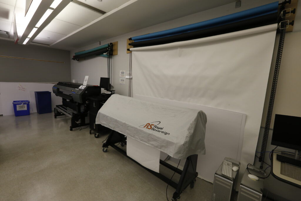 A brightly lit print studio or workshop featuring two large-format printers, one covered with a Royal Sovereign sheet, and multiple photography backdrop rolls (white, blue, black, turquoise) mounted on a wall. Several computer towers and a 'Vinyl Only' bin are also present, indicating a production-focused environment.