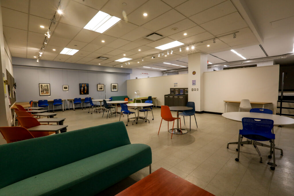 An inviting university lounge or common area featuring contemporary blue and orange chairs, green sofas, and diverse tables under bright lighting, with artwork on the walls.