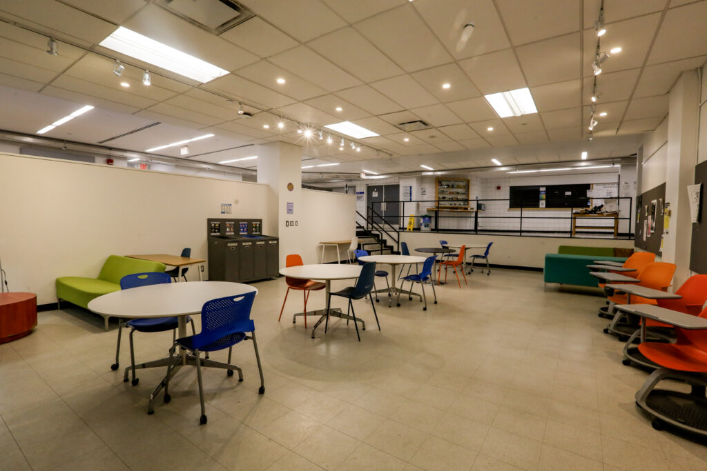 An open, modern common area or lounge, possibly in a school or office, with diverse seating arrangements including round tables with blue and orange chairs, and colorful sofas. A recycling station and staircase are visible in the background.