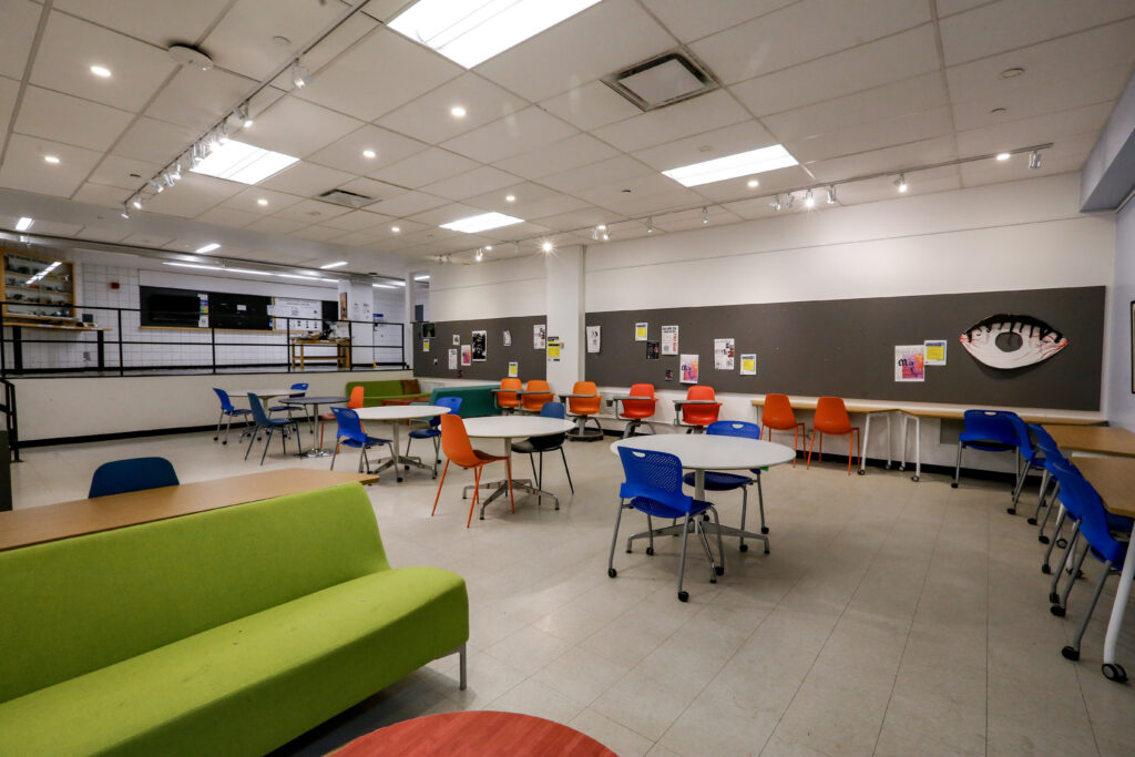 A spacious, modern common area or student lounge featuring circular white tables with bright blue, orange, and red chairs, lime-green seating in the foreground, and bulletin boards on the walls.