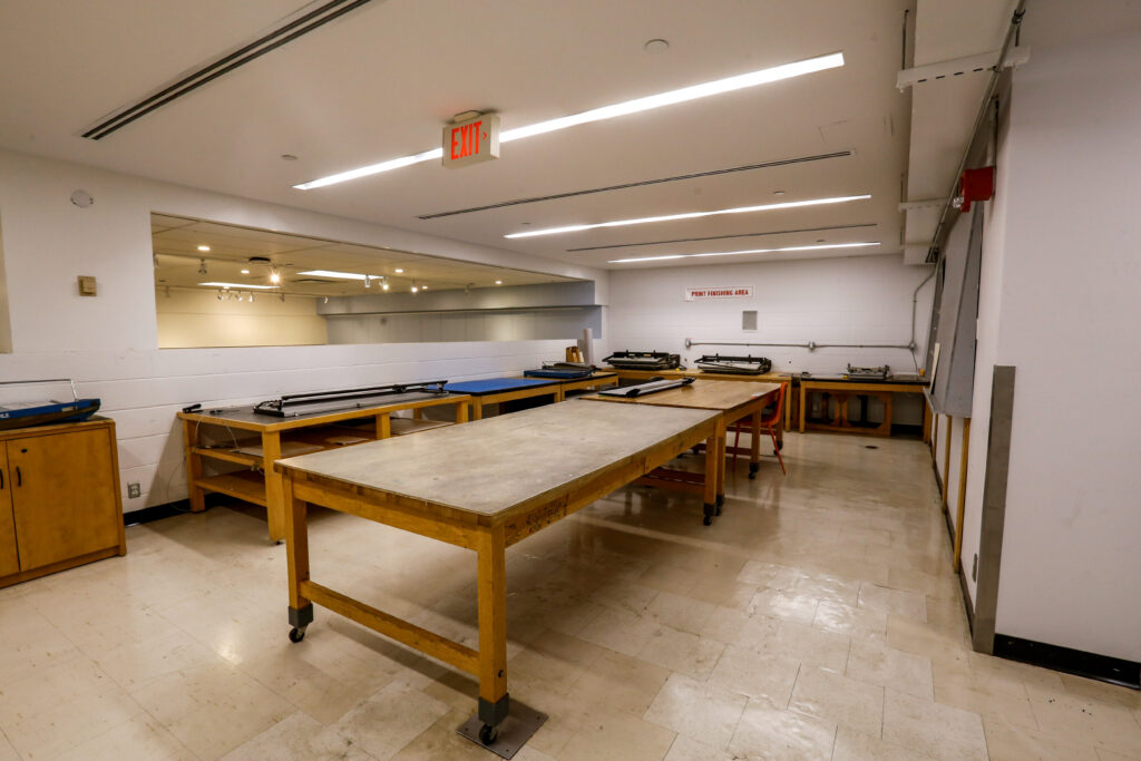 A well-lit print finishing area featuring several long wooden work tables, some with equipment, and a large mirror reflecting the space, with an 'EXIT' sign on the ceiling.