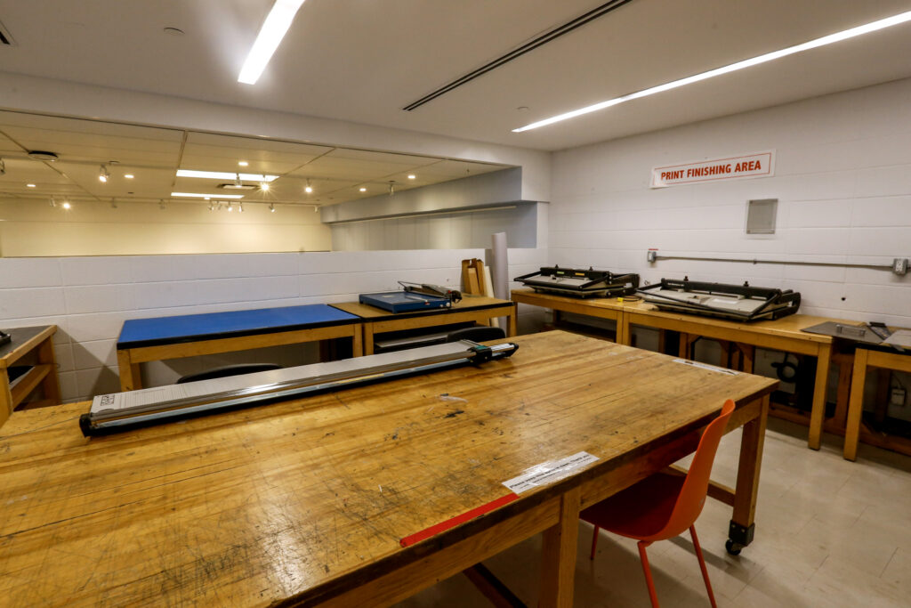 An empty print finishing area with a large wooden table in the foreground holding a paper trimmer, and other tables with finishing equipment along the walls, under a sign that reads 'PRINT FINISHING AREA'.