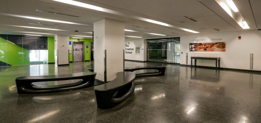 An interior view of a contemporary school hallway with polished dark floors, white walls, and bright green sections. Modern, black curved benches are in the foreground, with signs for 'The Creative School' and 'Image Arts Commons' visible.