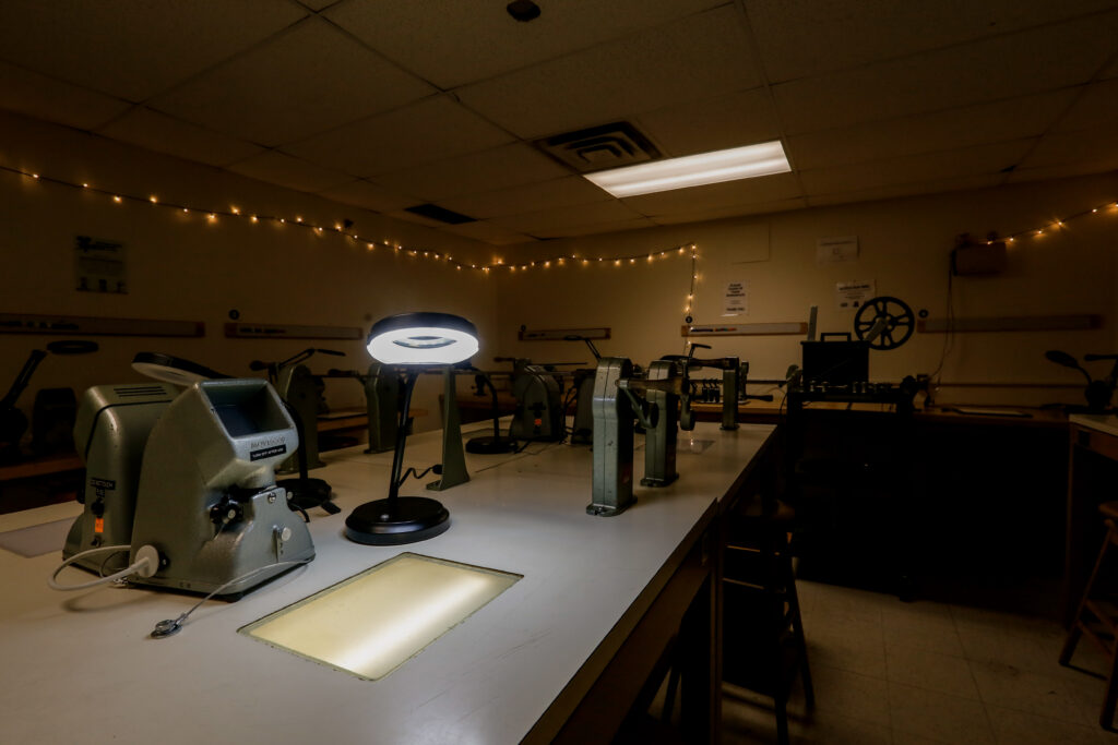 A specialized darkroom or film lab with multiple film editing machines, illuminated by a bright ring light, a light table, and decorative string lights.