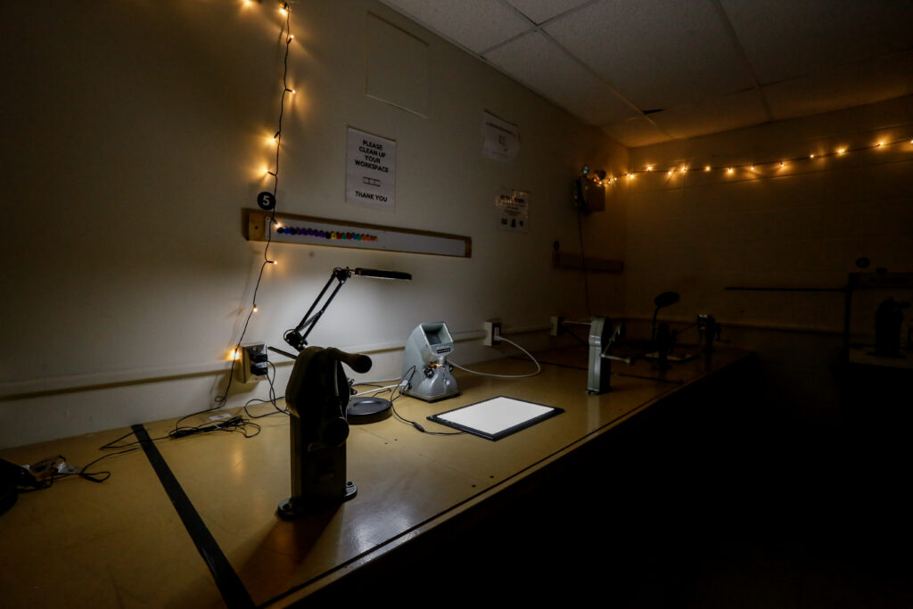 A retro film editing station in a dimly lit room, featuring a desk lamp, a Minolta film viewer, and winding equipment on a wooden bench, with string lights overhead.