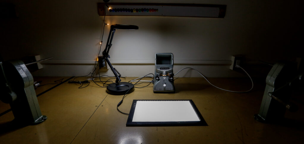A dimly lit film editing station features a Moviscop film viewer, an illuminated light pad, and an adjustable desk lamp, with large film reel mechanisms at either side.