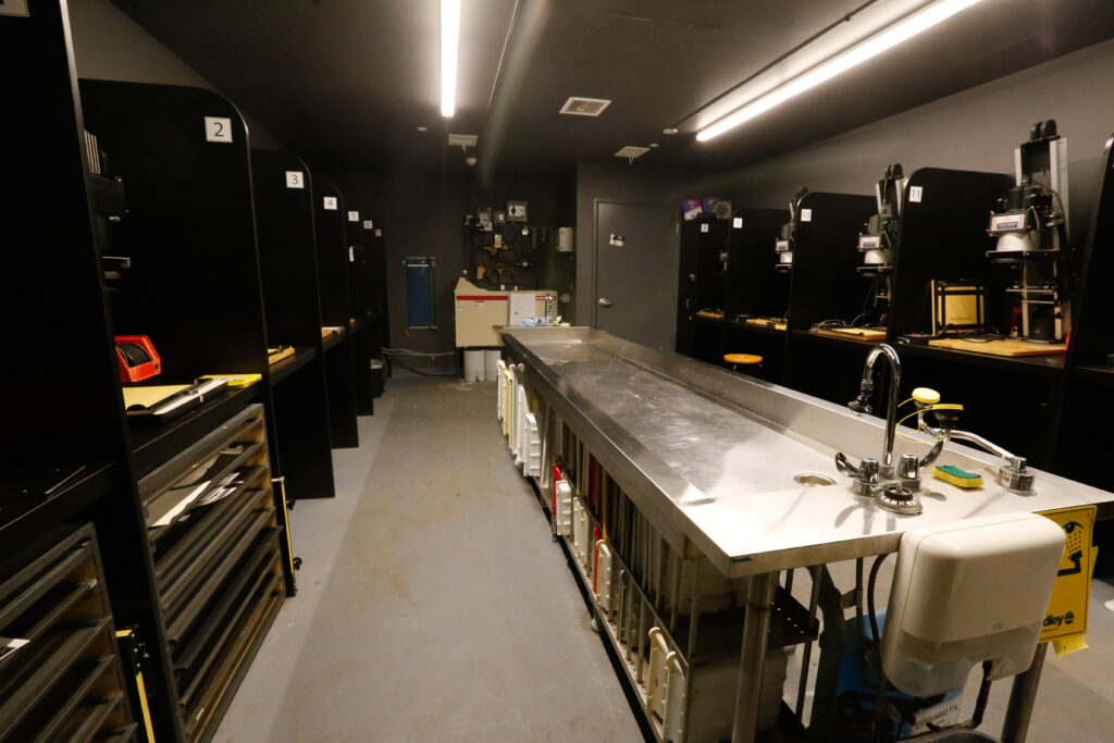 An empty photography darkroom featuring rows of individual enlarger workstations along the perimeter and a long stainless steel wet-processing counter with sinks and faucets in the center.