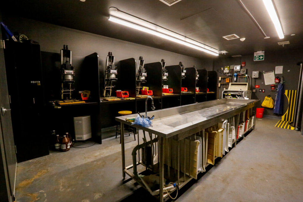 An operational photography darkroom featuring a long stainless steel developing sink in the foreground, with multiple enlarger stations lined against the back wall, and safety equipment visible on the right wall.