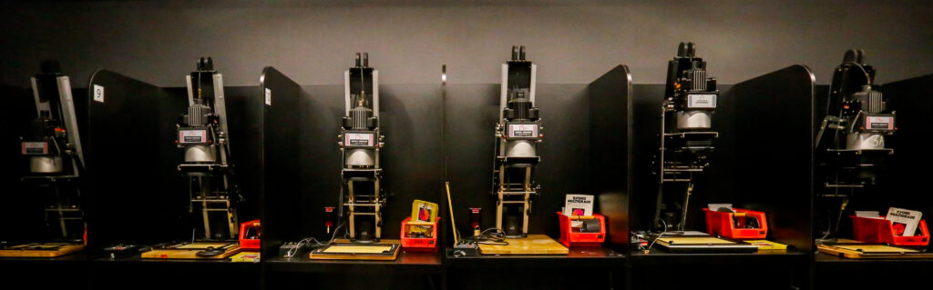 Six individual darkroom printing stations, each equipped with a photographic enlarger, paper safe, and timer, used for analog photo development.
