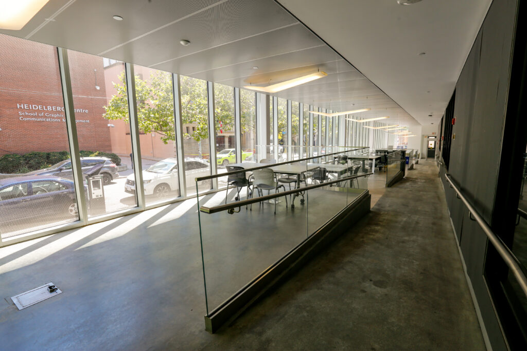 A contemporary study or common area in the Heidelberg Centre School of Graphic Communications Management, featuring multiple tables and chairs alongside large, sunlit windows that overlook an exterior campus scene with trees and cars.