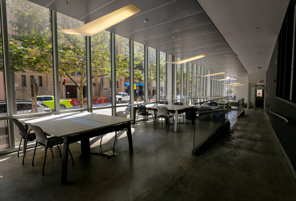 A bright, empty common area or library with floor-to-ceiling windows showcasing a busy city street with green trees, featuring long study tables and modern chairs.