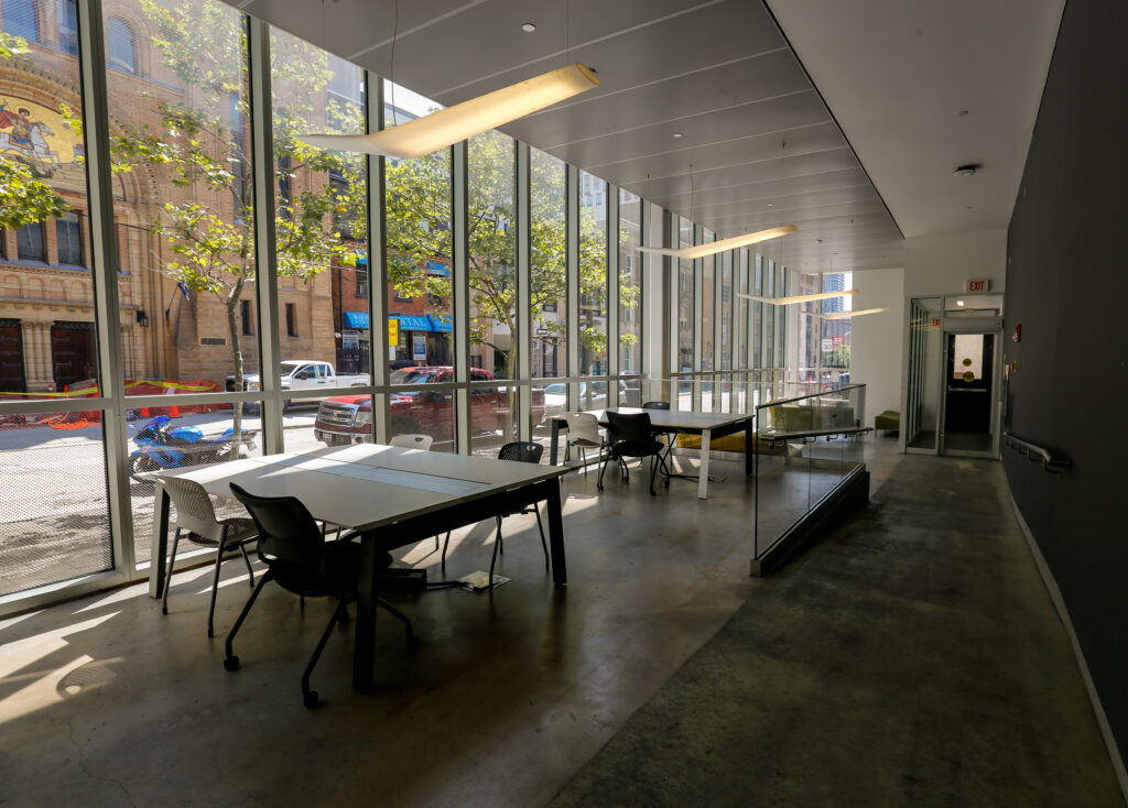 A bright, contemporary study or co-working space with concrete floors and large windows showing a bustling city street with a historic building and trees. Several tables and chairs are illuminated by both natural light and long, artistic pendant lamps.