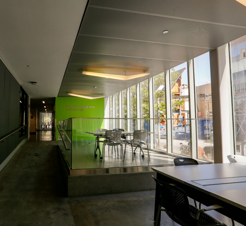 The School of Image Arts Student Lounge, featuring a vibrant green wall with the school's name, multiple tables and chairs on a raised platform, and large windows with a view of an urban area and construction. A long hallway extends to the left.