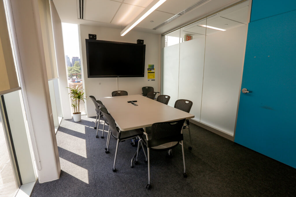 A bright, modern meeting room with a white table, eight black chairs, and a large TV screen. Windows on the left show a city view and cast sunlight shadows on the gray carpet, while a frosted glass partition and a blue door are on the right.