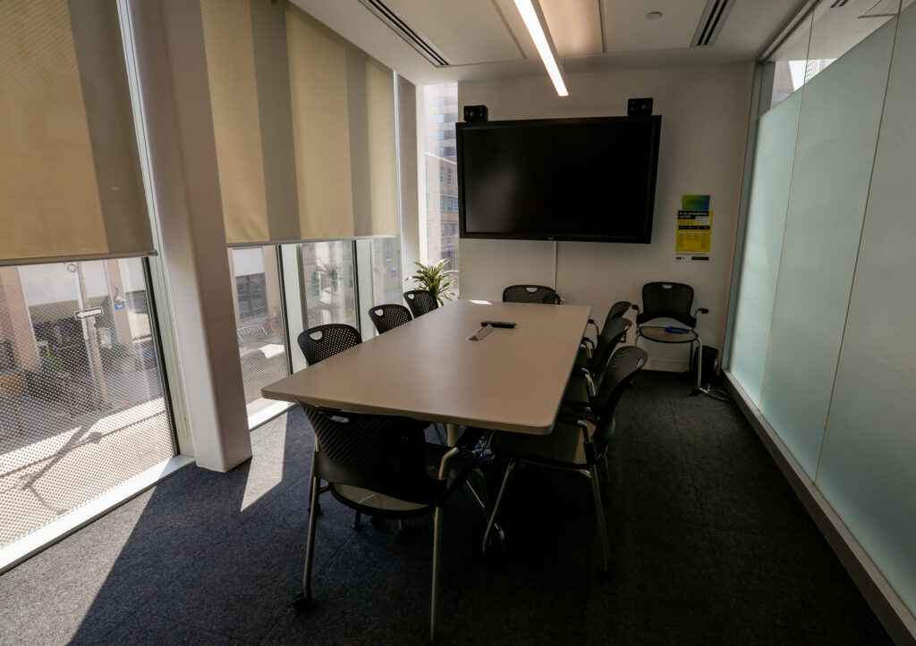 An empty conference room featuring a light-colored rectangular table with eight black chairs, a large wall-mounted TV with speakers, and large windows with partial beige blinds showing an urban view.