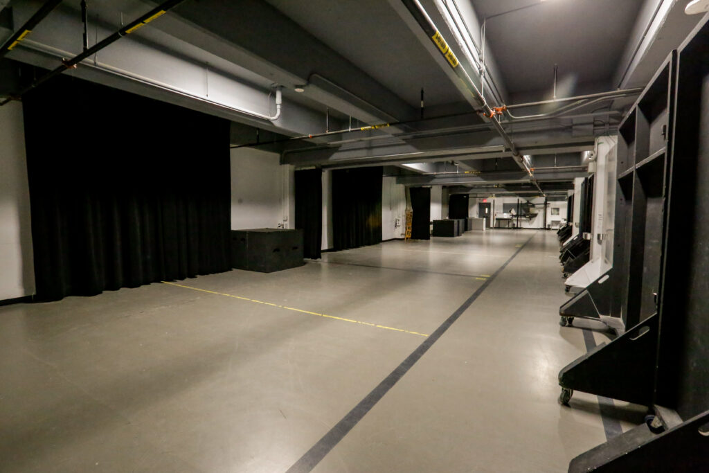 A long, unoccupied multi-purpose room or production studio featuring black stage curtains, an industrial ceiling with exposed pipes, a concrete floor with yellow and black lines, and a row of tall, wheeled scenic flats.