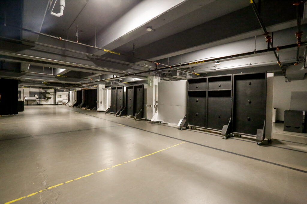 An empty, brightly lit studio or industrial space featuring a row of large, wheeled black and white soundproofing or partition panels on the right, with exposed pipes and ducts on the ceiling.