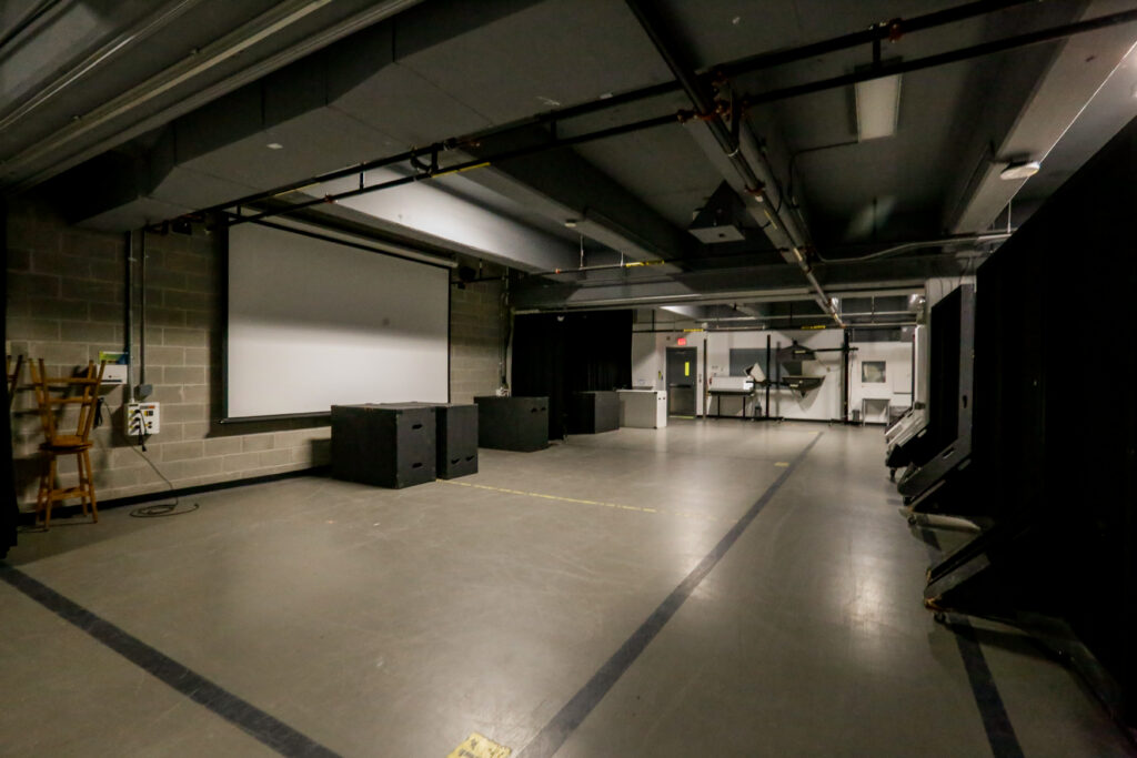 A dim, empty media studio or lecture hall featuring a large white projector screen on a cinder block wall, black stage boxes, and exposed ceiling utilities. A row of dark, movable panels lines the right side, with workstations visible in the background.
