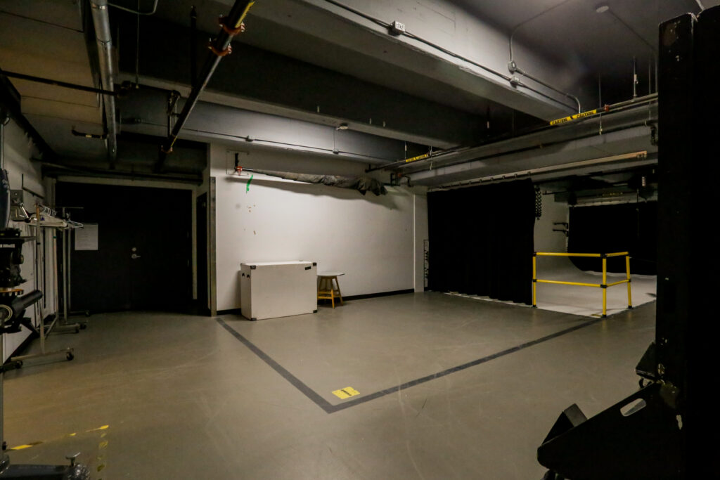 An industrial photography studio featuring a white seamless backdrop section, a large black curtain, various studio equipment, and exposed pipes and conduits on the ceiling.