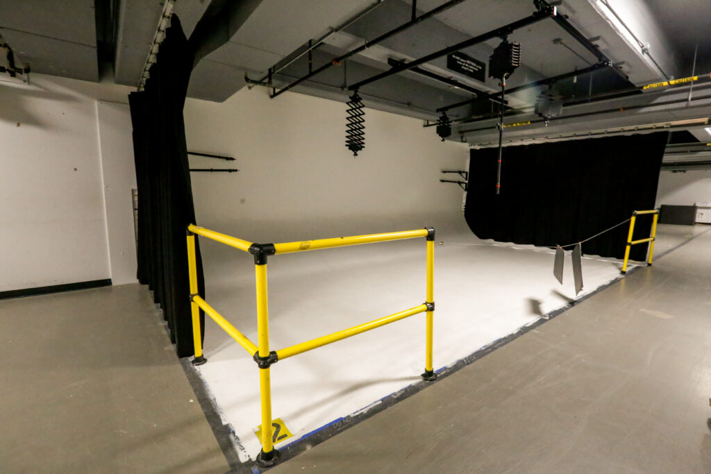 An empty professional photo and video studio setup, featuring a large white cyclorama, black curtains, yellow safety barriers, and overhead lighting equipment.