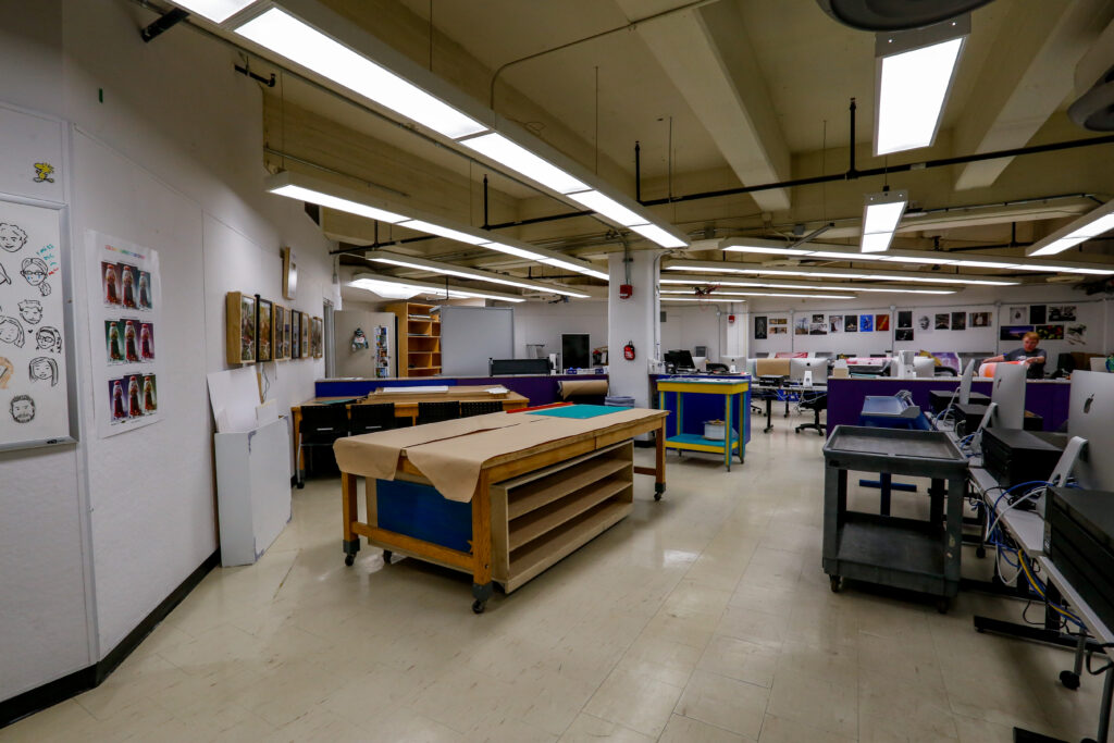 An active art studio or digital lab featuring multiple computer workstations, large worktables, and various art projects displayed on the walls, suggesting a creative learning or production environment.