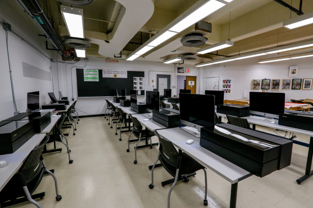An empty digital printing facility classroom with multiple workstations, each equipped with an Apple iMac computer and a large Epson printer, arranged in rows under bright fluorescent lights.