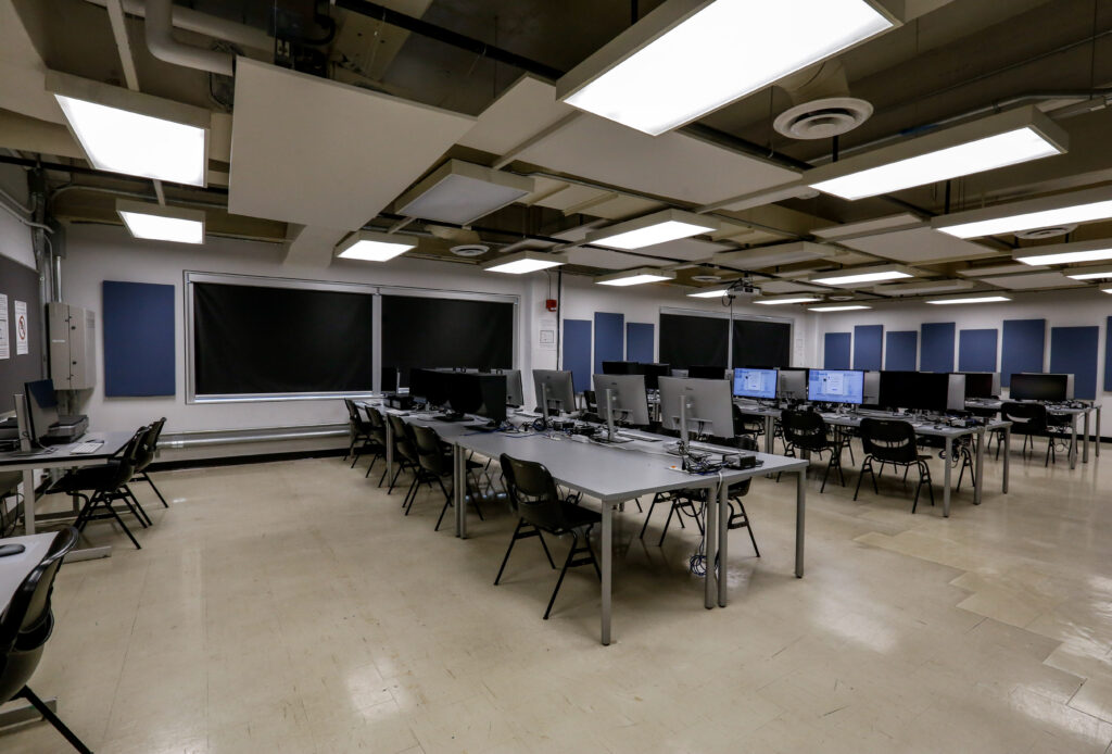 Unoccupied modern computer lab with rows of grey tables, silver desktop monitors, black chairs, and blue acoustic panels under an industrial ceiling.