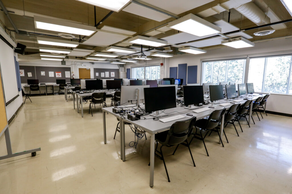 A modern computer lab with multiple workstations, featuring grey desks, black monitors, and chairs, under bright ceiling lights. Large windows show trees outside, and the room appears ready for use.