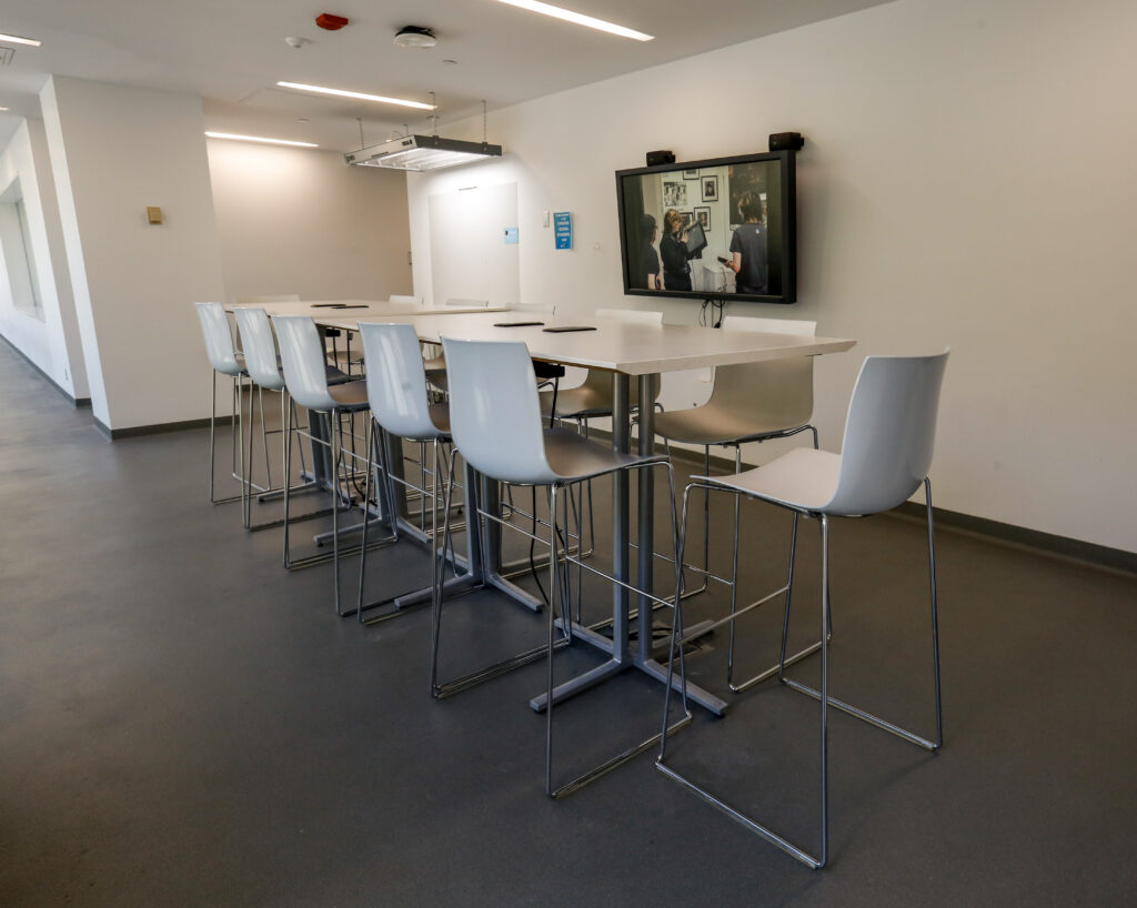 A contemporary university or office common area featuring several high-top tables with matching bar stools, and a large TV screen displaying a creative project, suggesting a collaborative and tech-equipped space.