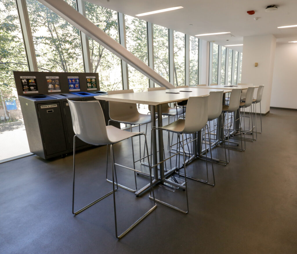 A bright, modern study or common area featuring a long high-top table with light grey bar stools, a multi-bin recycling station, and large windows offering a view of green trees.