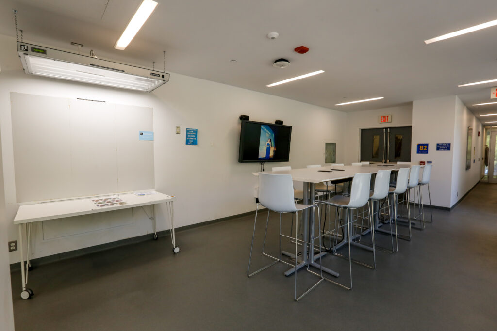 A well-lit university common area features a long white high-top table with eight gray bar stools, a large TV displaying an image, and a white presentation board with a specialized 'GTI Graphiclite' above it. An 'EXIT' sign is visible over a doorway on the right.