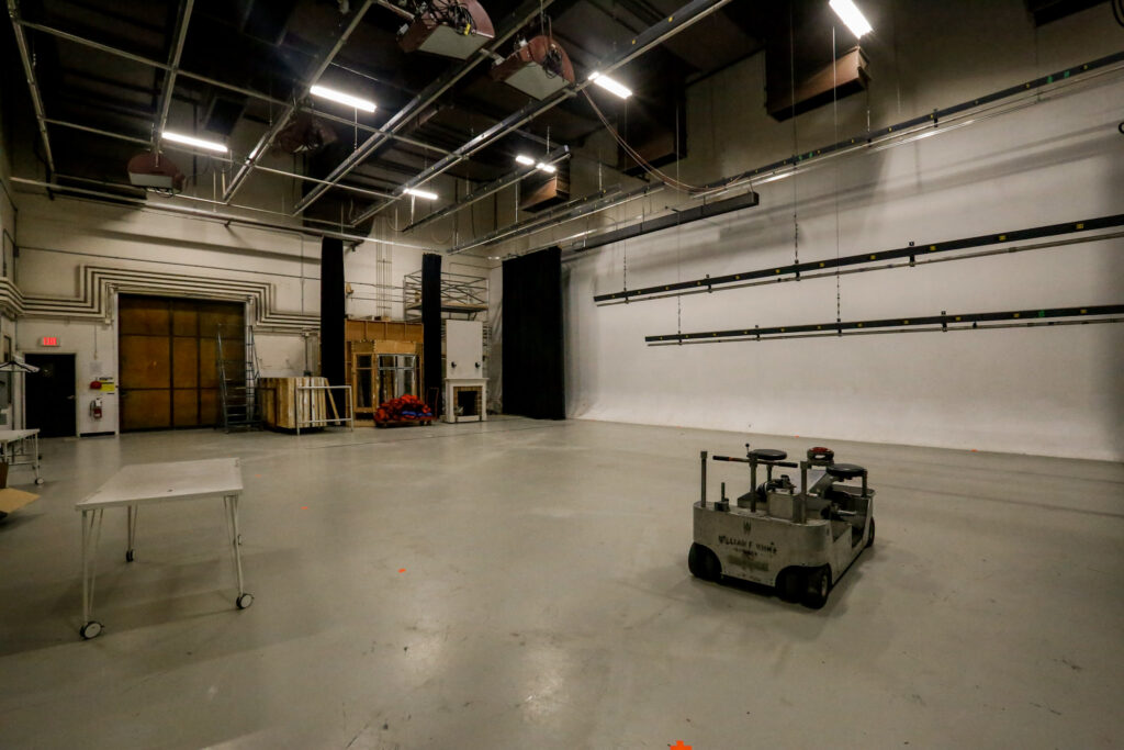 A large film or television studio showing a seamless white cyclorama, an overhead lighting grid, and a 'William F. White' production dolly, with various set pieces and equipment stored on the left.