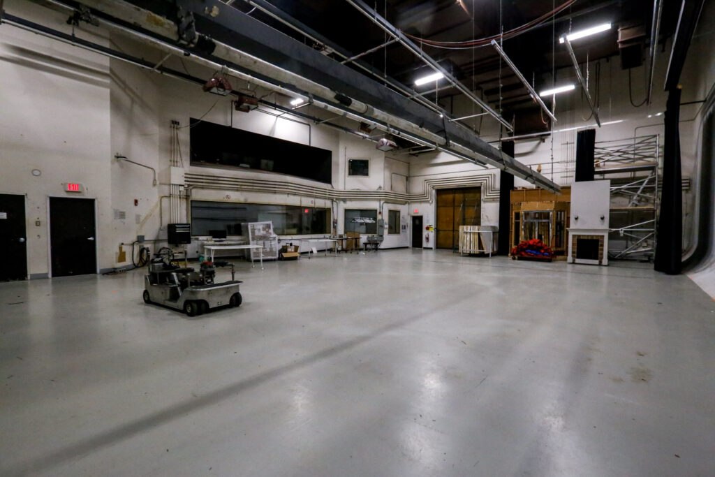 An expansive soundstage or workshop featuring a multi-wheeled robot in the foreground, an observation room window, and stage props like a fireplace and scaffolding in the background.
