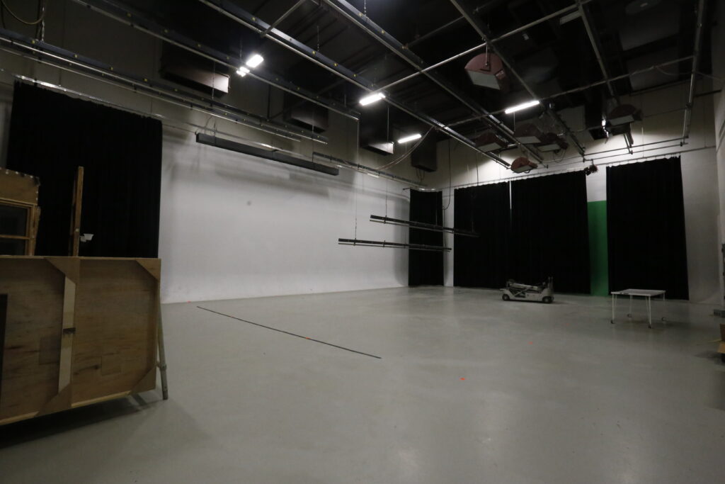 An unoccupied production studio ready for filming or photography, featuring a seamless white wall, black curtains, a green screen segment, and various lighting and production equipment.