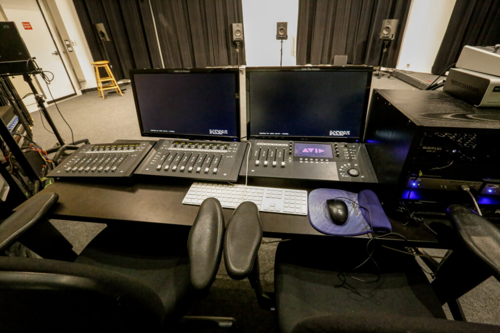 A studio control room setup with dual computer monitors, audio mixing consoles, and rack-mounted equipment, with studio monitor speakers and dark curtains in the background.
