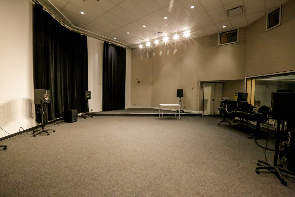 An empty, well-equipped audio studio featuring a stage with black curtains, multiple speakers, and a separate control room visible through a large window, under bright lighting.