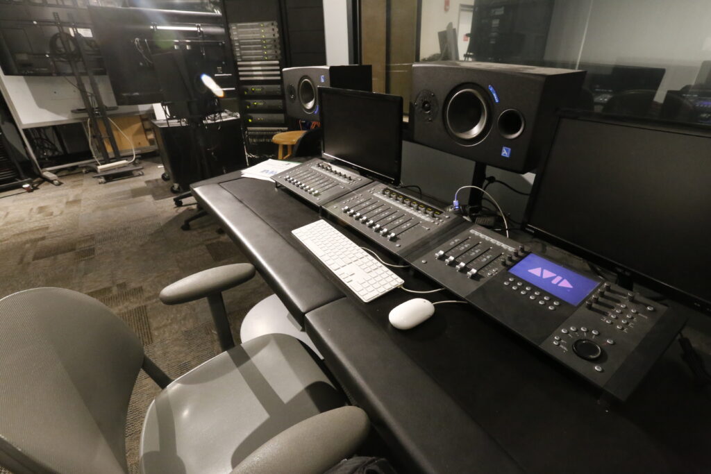 An Avid S1 control surface and dual studio monitors set up on a dark desk in a modern audio/video post-production studio, with server racks in the background.