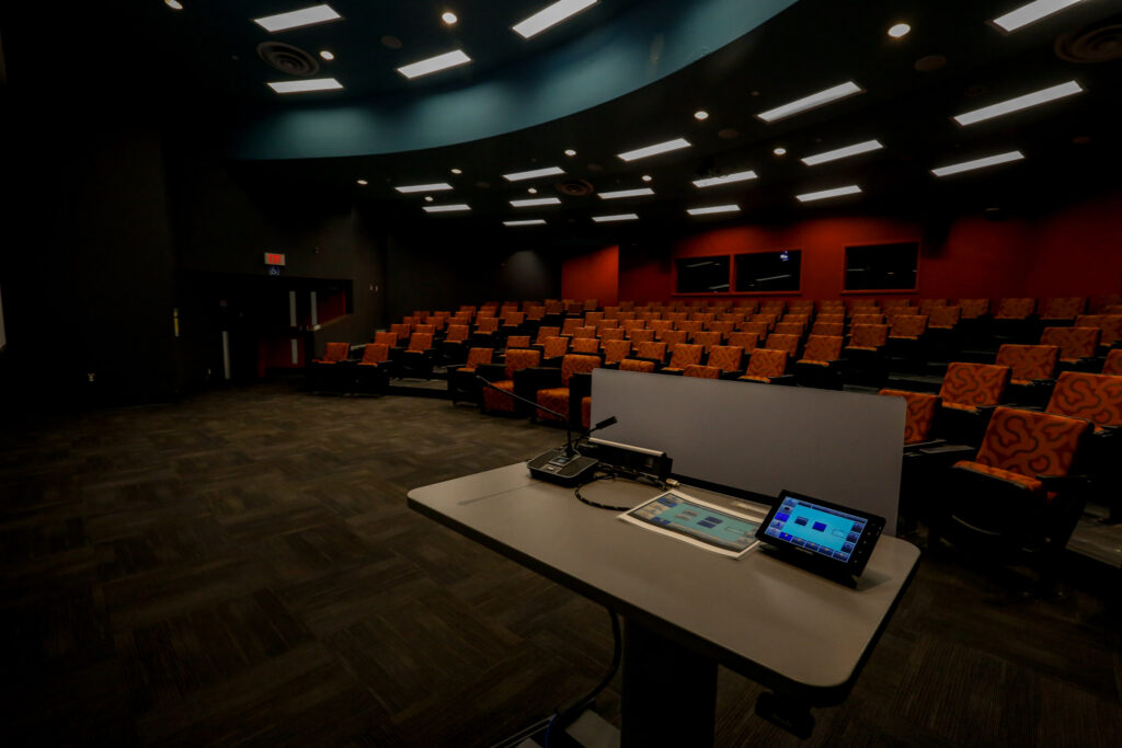 A contemporary, empty lecture hall equipped with an advanced lectern, featuring rows of orange and black patterned seats under bright overhead lighting, ready for a presentation.